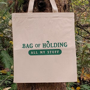Pode incluir: Uma sacola de lona bege com o texto "BAG OF HOLDING" e "ALL MY STUFF" impresso em verde. A sacola tem alças creme e está pendurada em um tronco de árvore, cercada por folhagens verdes.