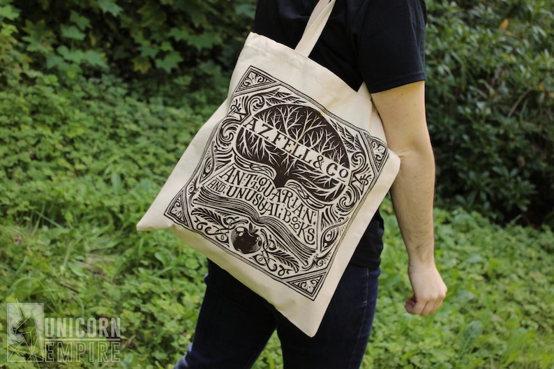 An off-white tote bag with black ink screen printing is worn over a model's shoulder. The text of the design reads AZ FEL & CO. Antiquarian and Unusual Books.