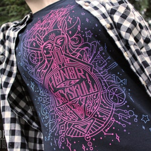 Puede incluir: Camiseta negra con un vibrante dise&ntilde;o gr&aacute;fico en rosa ne&oacute;n y azul. El dise&ntilde;o presenta un emblema circular con el texto "For the Honor of Skryll" y formas geom&eacute;tricas. Una camisa a cuadros en blanco y negro se usa encima.