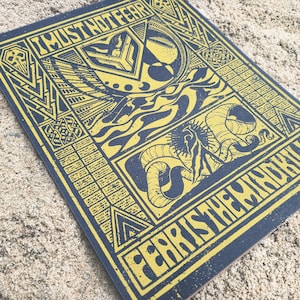 May include: A square, navy blue print with yellow text and illustrations. The top text reads "I MUST NOT FEAR", and the bottom text reads "FEAR IS THE MIND KILLER". The print features geometric shapes, a crescent moon, and a serpent.