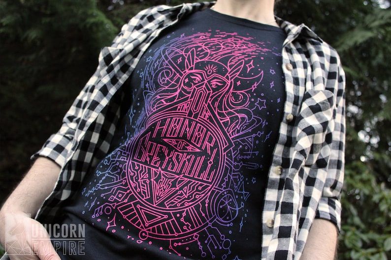 Puede incluir: Camiseta negra con un dise&ntilde;o gr&aacute;fico rosa ne&oacute;n y azul con el texto "HONOR OF GREYSKULL". El dise&ntilde;o incluye una cara estilizada, formas geom&eacute;tricas y estrellas. Una camisa a cuadros en blanco y negro se usa encima.