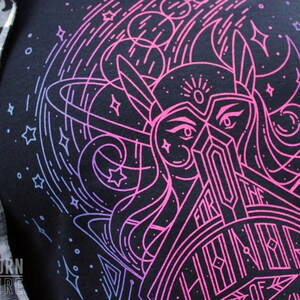 Puede incluir: Camiseta azul marino oscuro con un gr&aacute;fico rosa ne&oacute;n y azul de una mujer guerrera con el texto "FOR THE HONOR OF". El dise&ntilde;o incluye estrellas y elementos celestiales. La camiseta est&aacute; parcialmente cubierta por una camisa a cuadros en blanco y negro.