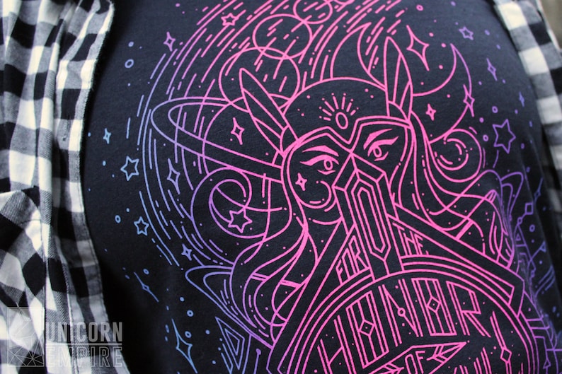 Puede incluir: Camiseta azul marino oscuro con un gr&aacute;fico rosa ne&oacute;n y azul de una mujer guerrera con el texto "FOR THE HONOR OF". El dise&ntilde;o incluye estrellas y elementos celestiales. La camiseta est&aacute; parcialmente cubierta por una camisa a cuadros en blanco y negro.