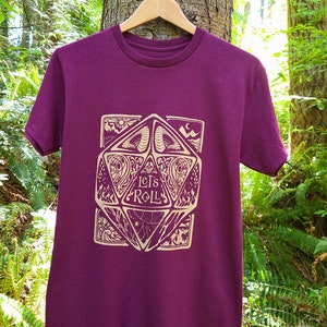 May include: A maroon t-shirt featuring a gold graphic of a 20-sided die with the words "Let's Roll" inside. The die is surrounded by intricate designs.