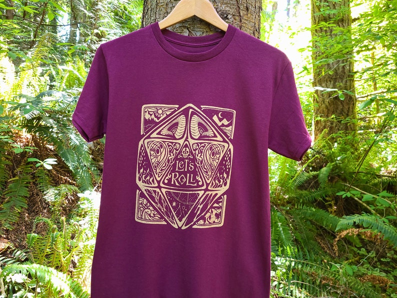 May include: A maroon t-shirt featuring a gold graphic of a 20-sided die with the words "Let's Roll" inside. The die is surrounded by intricate designs.