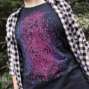 Puede incluir: Camiseta negra con un dise&ntilde;o gr&aacute;fico rosa y azul ne&oacute;n que incluye el texto "Honor of Greyskull". La camiseta se usa con una camisa a cuadros en blanco y negro.