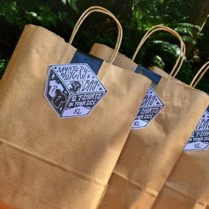 May include: Several brown paper shopping bags with white handles are displayed. Each bag has a sticker that reads "MYSTERY BAG" with the text "2 T-SHIRTS IN YOUR SIZE XL". The bags are arranged in a row, with a green leafy background.