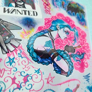 May include: A sticker sheet featuring a colorful illustration of a female character with blue hair and a pink background. The character is holding a gun and has a cloud-like design on her arm. The sticker sheet also includes a rocket, stars, and the word "WANTED".
