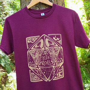 May include: Burgundy t-shirt featuring a gold graphic of a dice with the words "Let's Roll" on it.