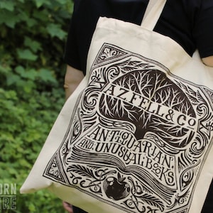 A closeup of the AZ FEL & CO tote bag shows more details. The design looks like the logo of a book store with an open book, feathers, and an apple.