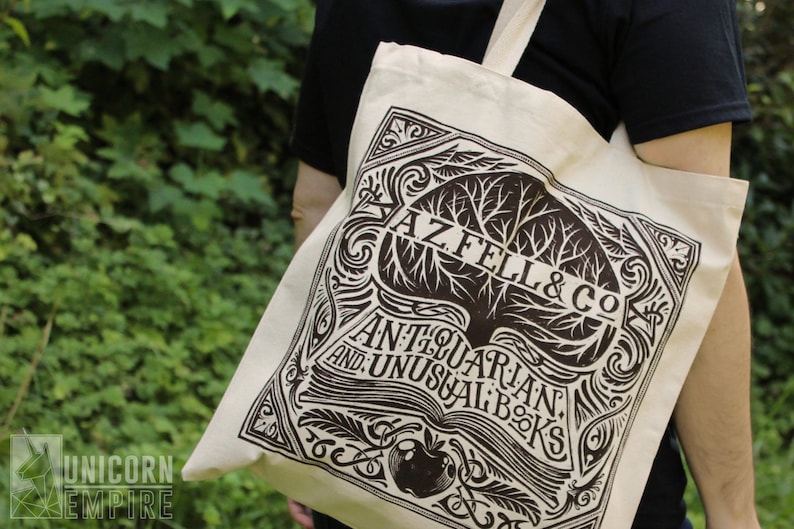 A closeup of the AZ FEL & CO tote bag shows more details. The design looks like the logo of a book store with an open book, feathers, and an apple.