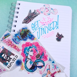 May include: A collection of stickers featuring a cartoon character with blue hair and a pink and blue cloud background. The stickers also include a rocket ship, a cloud, and a character with a gun. The text "Get Jinxed!" is written in blue and pink on a white notebook.
