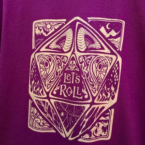 May include: Purple t-shirt featuring a gold graphic of a 20-sided die with the text "Let's Roll".