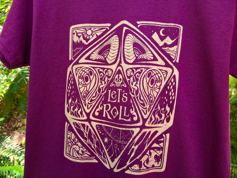 May include: Purple t-shirt featuring a gold graphic of a 20-sided die with the text "Let's Roll".