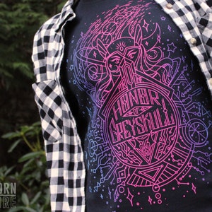 Puede incluir: Camiseta negra con un dise&ntilde;o gr&aacute;fico rosa y azul vibrante con el texto "Honor of Greyskull". El dise&ntilde;o incluye formas geom&eacute;tricas y un casco estilizado. La persona lleva una camisa a cuadros en blanco y negro.