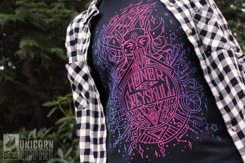 Puede incluir: Camiseta negra con un dise&ntilde;o gr&aacute;fico rosa y azul vibrante con el texto "Honor of Greyskull". El dise&ntilde;o incluye formas geom&eacute;tricas y un casco estilizado. La persona lleva una camisa a cuadros en blanco y negro.