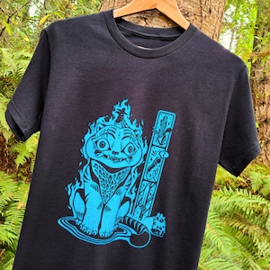 May include: Black t-shirt featuring a blue illustration of a cat engulfed in flames, with a matching blue design on a vertical rectangle. The cat has a surprised expression. The shirt is hanging on a wooden hanger against a natural background.
