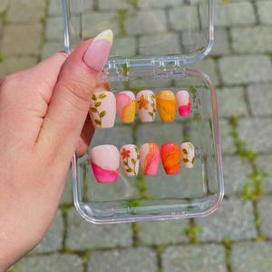 May include: A set of press-on nails in a clear plastic case. The nails feature various designs, including floral patterns and abstract colour gradients in shades of pink, orange, and yellow. Some nails have a French tip design.