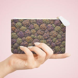 May include: A rectangular zippered pouch with a colorful pattern of small, round shapes in shades of green, purple, and orange. The pouch has a white zipper pull and a gold-colored zipper. The pouch is held in a hand against a pink background.
