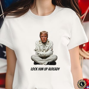 May include: White t-shirt featuring a seated figure in a straitjacket. The image includes the text "LOCK HIM UP ALREADY" in black font. The background is partially obscured by the shirt.