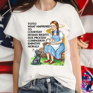 May include: White t-shirt featuring a graphic of Dorothy from The Wizard of Oz with Toto. The text reads "TOTO WHAT HAPPENED TO COURTESY HUMAN RIGHTS DUE PROCESS COMPASSION EMPATHY MORALS ?". Dorothy is wearing a blue and white checkered dress.