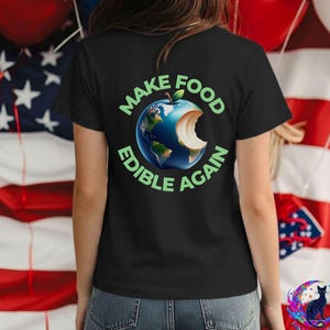May include: A black short-sleeved t-shirt featuring a graphic on the back. The design shows a blue and green Earth shaped like an apple with a bite taken out of it. The text "MAKE FOOD EDIBLE AGAIN" is printed in light green capital letters.