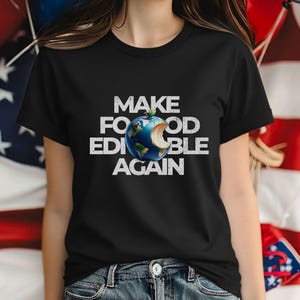 May include: A black short-sleeved t-shirt featuring white distressed text that reads MAKE FOOD EDIBLE AGAIN. In the center, a graphic depicts a blue and green Earth shaped like an apple with a bite taken out of the side, showing a white core.