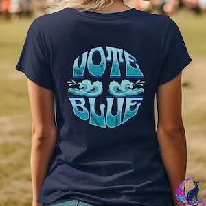 May include: A dark navy blue short-sleeve t-shirt featuring a circular graphic on the back. The graphic contains the text VOTE BLUE in a stylized light blue font, separated by two illustrated ocean waves in shades of light and dark blue.
