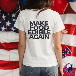 May include: A white short-sleeved t-shirt featuring the black text MAKE FOOD EDIBLE AGAIN printed in a bold, sans-serif font on the back. The shirt has a crew neckline and a standard fit, showcasing a clean design against a patriotic background.