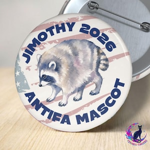 Op de afbeelding: Een ronde pin-back button met een aquarelillustratie van een wasbeer in het midden. De tekst JIMOTHY 2026 ANTIFA MASCOT is in donkerblauwe hoofdletters rondom de wasbeer gedrukt, op een achtergrond met vervaagde rode en witte strepen.