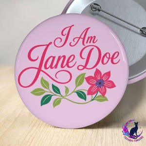 May include: A round pinback button with a light pink background featuring the dark pink cursive text I Am Jane Doe. Below the text is a graphic of a single pink flower with a dark purple center and green leaves on a curved stem. The back is silver metal.