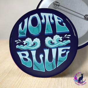 May include: A circular pin-back button featuring a dark blue background with the text VOTE BLUE in a light blue, stylized font. Two light blue, graphic wave illustrations are positioned between the words VOTE and BLUE on the button surface.