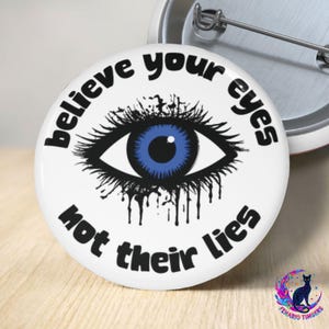 May include: White button with the text "believe your eyes not their lies" encircling a blue eye graphic. The eye has black eyelashes and a black outline. A silver pin is attached to the back.
