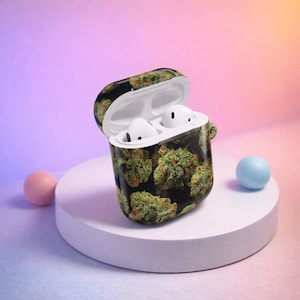 May include: An open black and green patterned AirPod case with white AirPods inside. The case is decorated with a green and brown cannabis bud design. The case is sitting on a white circular platform with a pink and blue ball on either side.