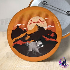 Op de afbeelding: Een ronde pin-back button met een grafische illustratie van een grijze wasbeer die voor donkerbruine bergen loopt tegen een zonsondergangachtergrond met oranje horizontale lijnen. De naam Jimothy staat in een schrijfletter boven de bergen.