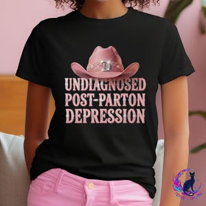 May include: A black short-sleeved t-shirt featuring a graphic of a pink cowboy hat with a sparkling band. Below the hat, the text reads UNDIAGNOSED POST-PARTON DEPRESSION in pink, glittery, block-style lettering with small sparkling accents.
