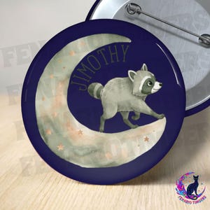 Op de afbeelding: Een ronde button met speldsluiting op een donkerblauwe achtergrond met een aquarel-stijl halve maan in zachte groen- en oranjetinten. Een grijze wasbeer loopt over de maan, met de naam JIMOTHY gedrukt in donkergroene letters.