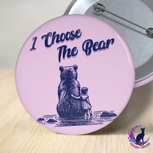 May include: A round pinback button with a light pink background featuring a dark blue illustration of a person standing next to a large bear. The text "I Choose The Bear" is printed in dark blue above the illustration. The back has a metal pin.