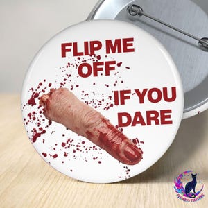 May include: A round pinback button with a white background featuring a severed human finger graphic covered in red blood splatters. The text FLIP ME OFF IF YOU DARE is printed in bold red capital letters across the surface of the button.
