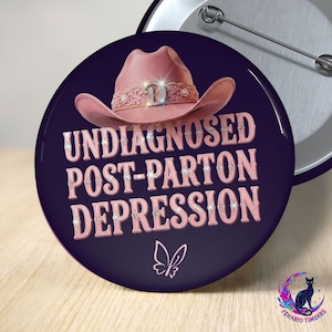 May include: A circular pin-back button with a dark purple background. It features a pink cowboy hat with a rhinestone band at the top. Below the hat, the text reads UNDIAGNOSED POST-PARTON DEPRESSION in pink letters with rhinestone accents.