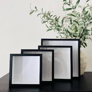 May include: Four black shadow box frames of varying sizes are arranged on a dark surface. Each frame has a white interior. A vase with green and white leafy branches is in the background, adding a touch of nature to the display.