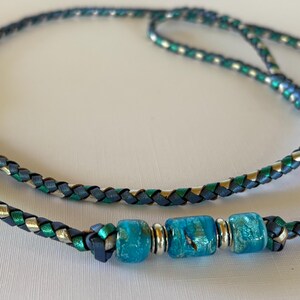 May include: A braided necklace featuring a cord woven from dark blue, metallic green, and silver strands. The center of the necklace is adorned with three textured, translucent turquoise glass beads separated by small, polished silver-toned metal spacers.