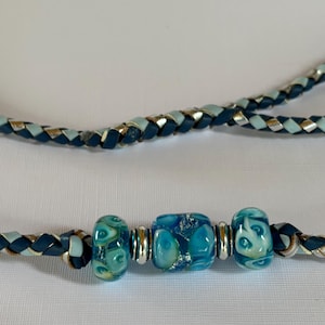 May include: A braided cord necklace featuring a combination of dark blue, light blue, and metallic silver strands. The center of the cord is adorned with three glass beads in shades of turquoise and blue, separated by small silver-toned metal spacers.