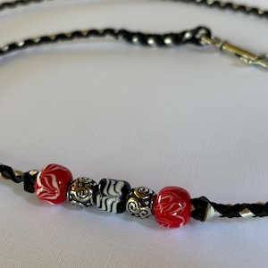 May include: A black braided leather leash featuring a silver metal swivel snap hook and a decorative section of five beads, including two red marbled glass beads, two silver-toned metal beads with etched swirls, and one black and white striped bead.