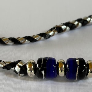 May include: A braided black and silver necklace with two dark blue glass beads and two black glass beads. The necklace has a silver clasp and is displayed on a white surface.