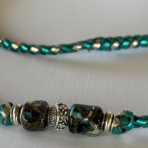 May include: A braided necklace featuring a metallic teal and silver cord. The center is adorned with two dark, marbled glass beads in shades of teal and black, separated by a small, textured silver-toned metal spacer bead and flanked by silver rings.