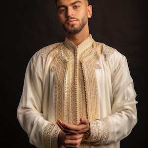 May include: A cream-coloured, long-sleeved garment with detailed gold embroidery. The garment has intricate patterns on the front, sleeves, and collar. The background is a solid black, which accentuates the ornate details of the clothing.