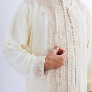 May include: A cream-coloured, long-sleeved garment with detailed beige embroidery. The embroidery adorns the collar, front, and cuffs. It features a high, mandarin-style collar and a button-up front. The fabric appears lightweight and flowing.