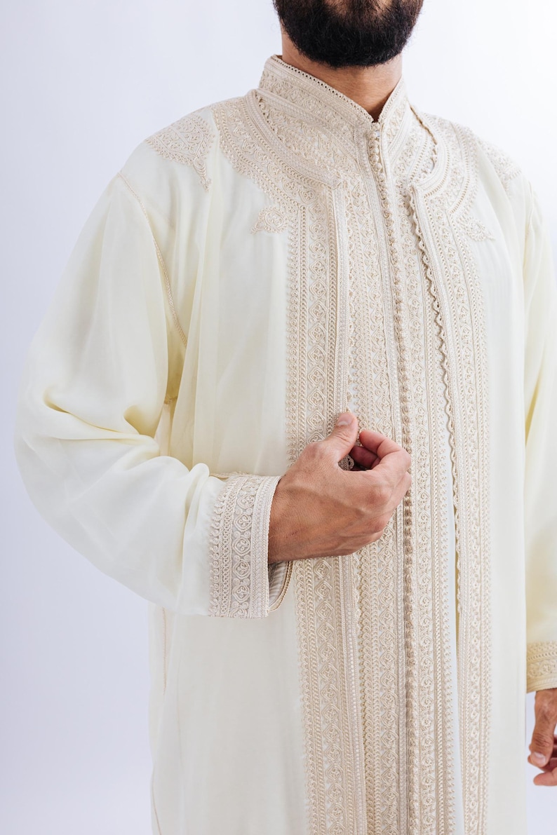 May include: A cream-coloured, long-sleeved garment with detailed beige embroidery. The embroidery adorns the collar, front, and cuffs. It features a high, mandarin-style collar and a button-up front. The fabric appears lightweight and flowing.
