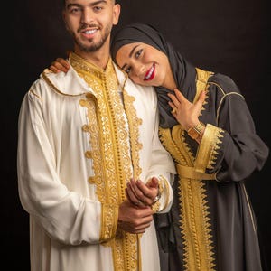 May include: A couple in traditional attire. The man wears a white robe with gold embroidery, while the woman wears a black dress with gold trim and a black hijab. The background is a solid black.